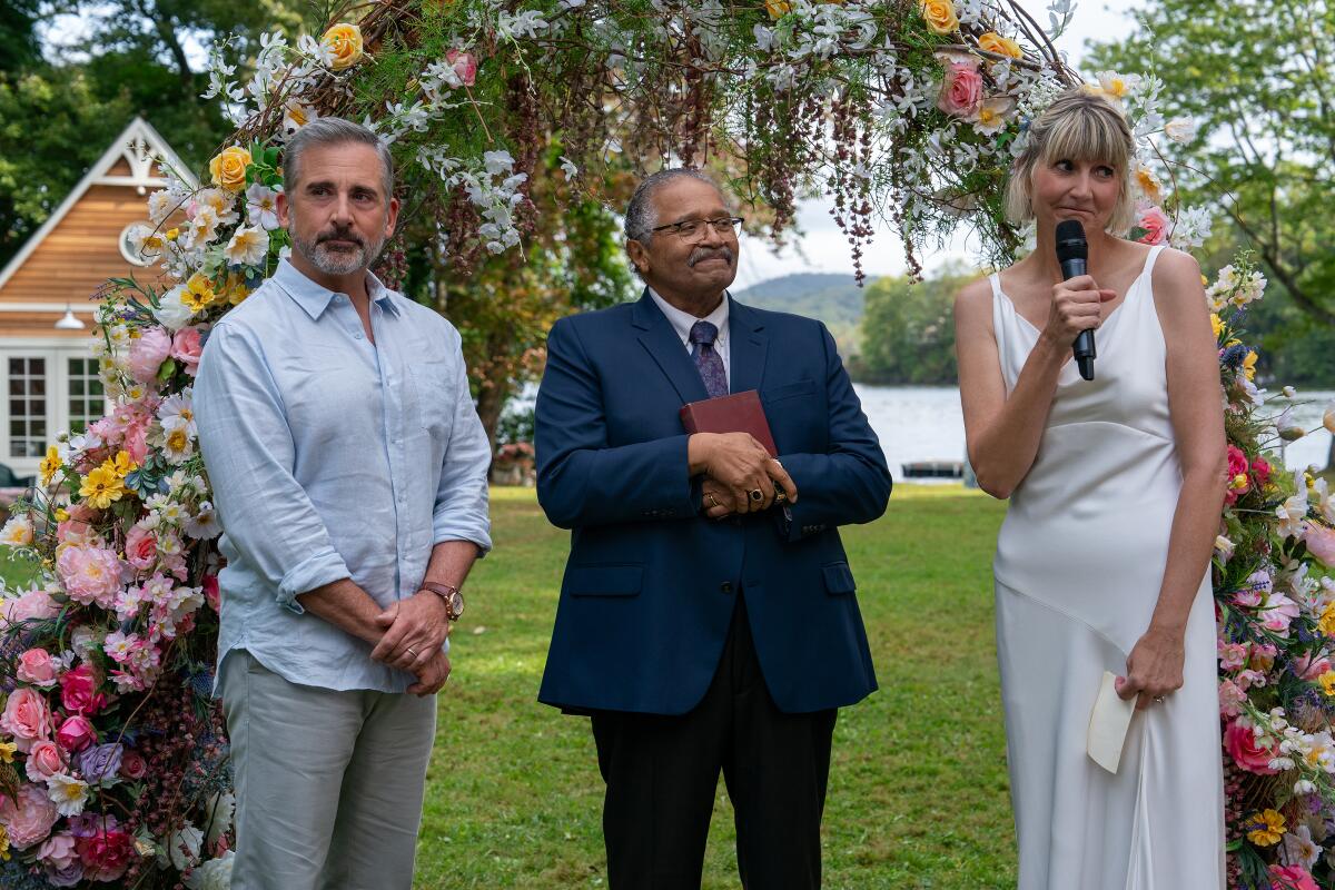 A couple stand near a wedding arch covered in flowers as an officiant in a blue suit stands under it with a book.