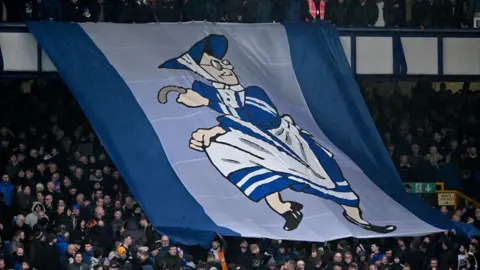 Getty Images Large flag at Goodison Park being passed over the heads of fans. It features a cartoon image of an old woman dressed in the Everton toffee lady uniform of a blue dress and white pinny.