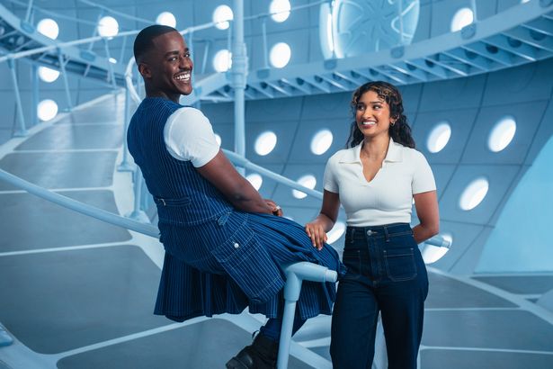 Ncuti Gatwa, in a blue outfit and white top, and Varada Sethu, in a white top and jeans, stood on the TARDIS set of Doctor Who in a promo photo.
