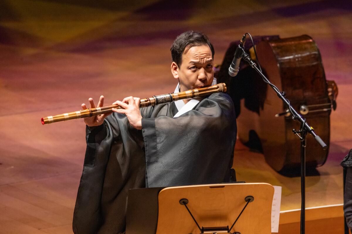 A man in traditional Korean clothing plays a daegeum, a large wooden flute.