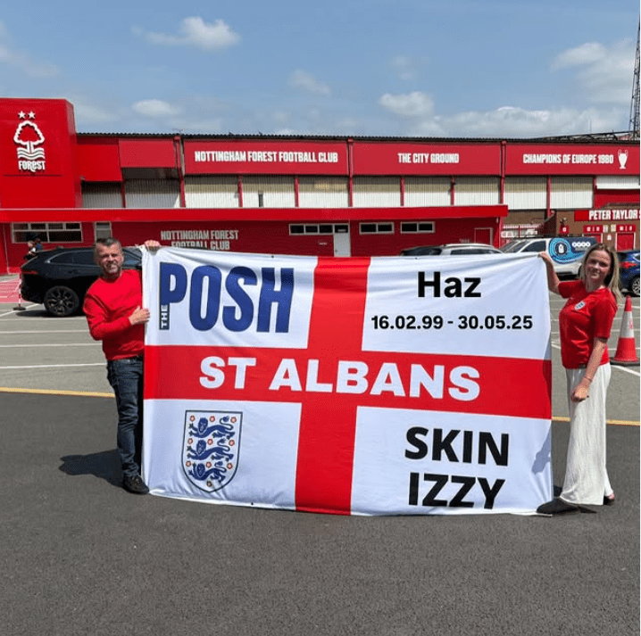 Two people holding a large St. Albans flag outside Nottingham Forest Football Club.