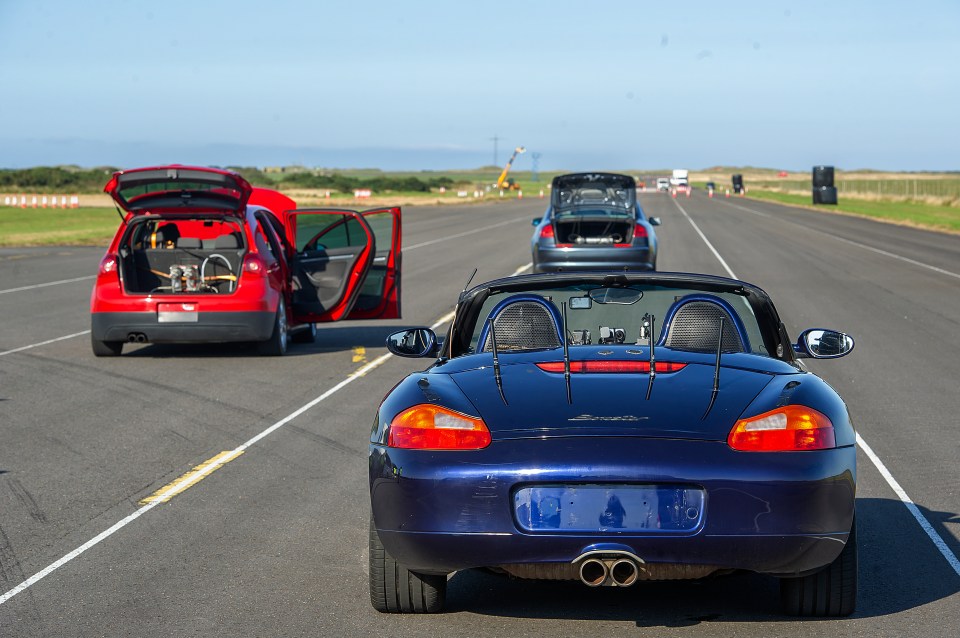 Rear view of a blue Porsche Boxster, a red Golf GTI, and a gray Audi on a race track.