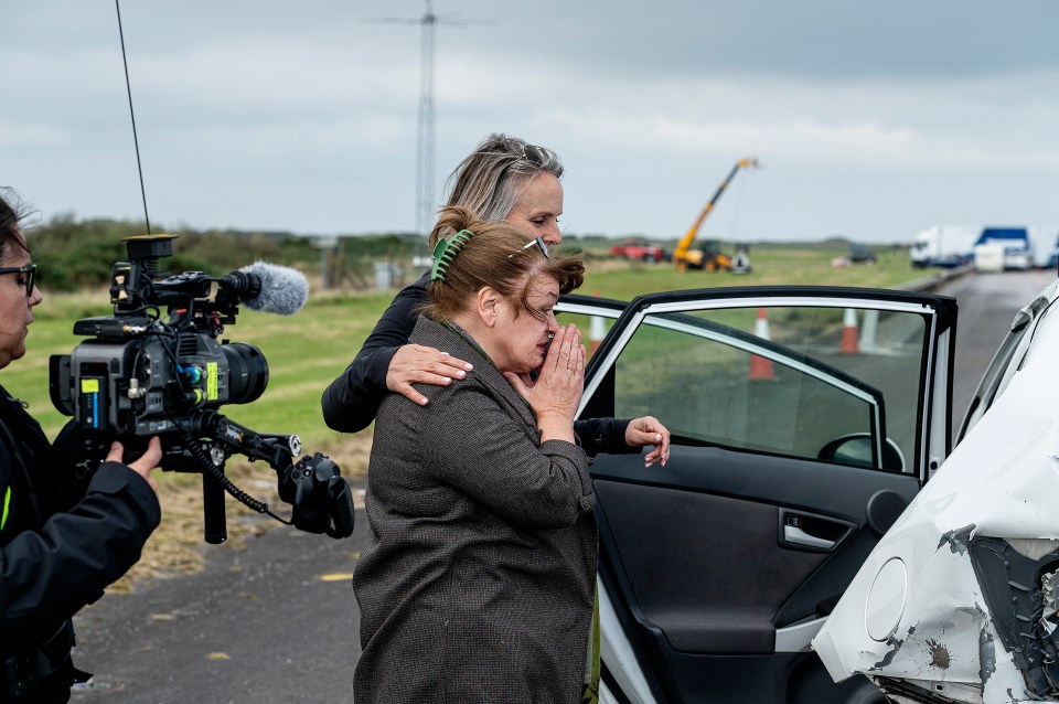 A woman comforts another woman next to a damaged car while a camera crew films.