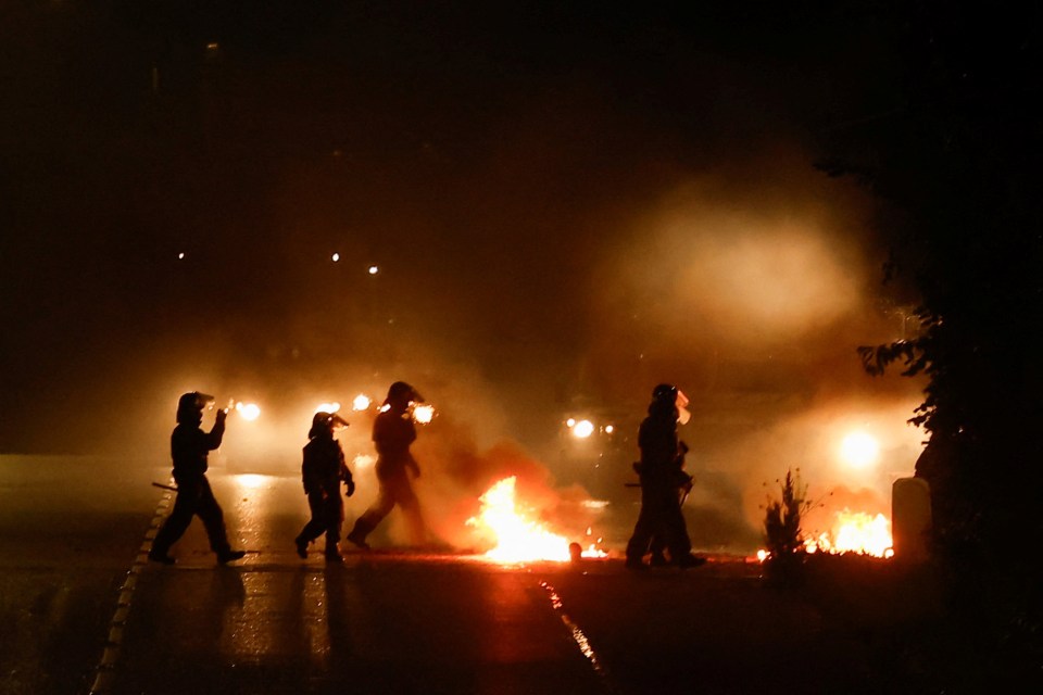 Riot police officers walking near fires during a protest at night.