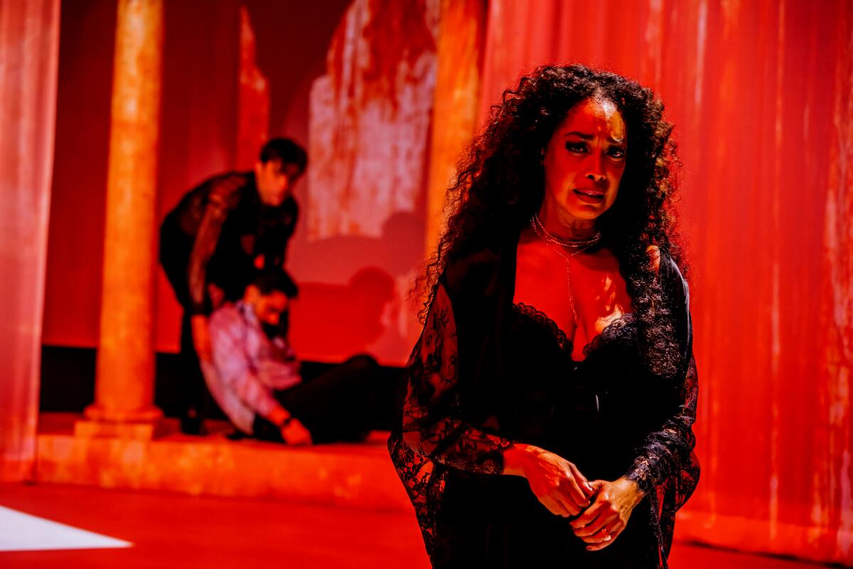 Bathed in red light, a woman stands as a man drags another man behind her in the play "Hamlet" at the Mark Taper Forum.