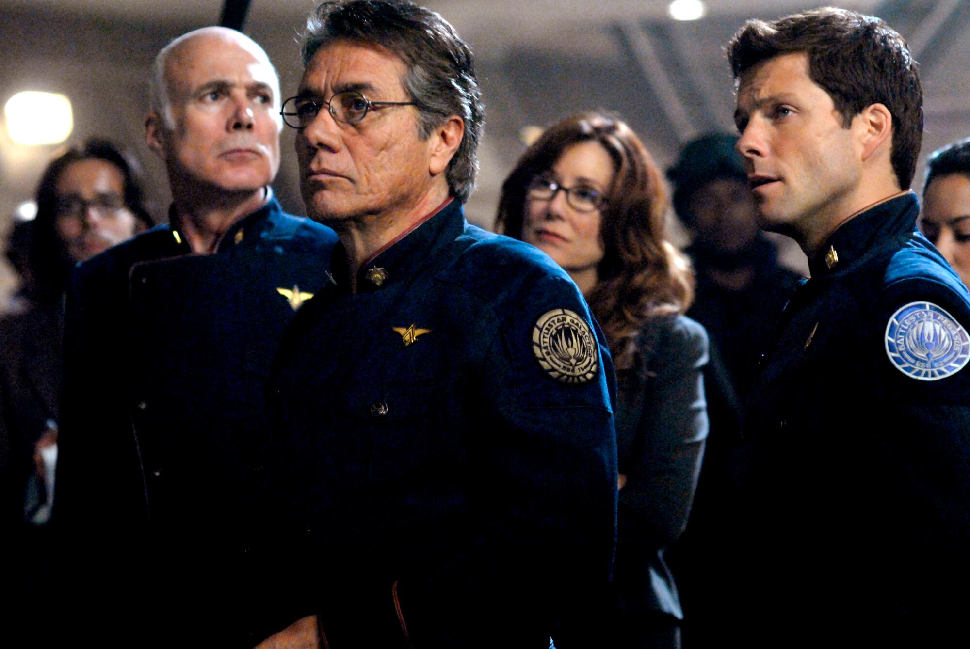 Three men and a woman in dark uniforms stand looking in different directions. 