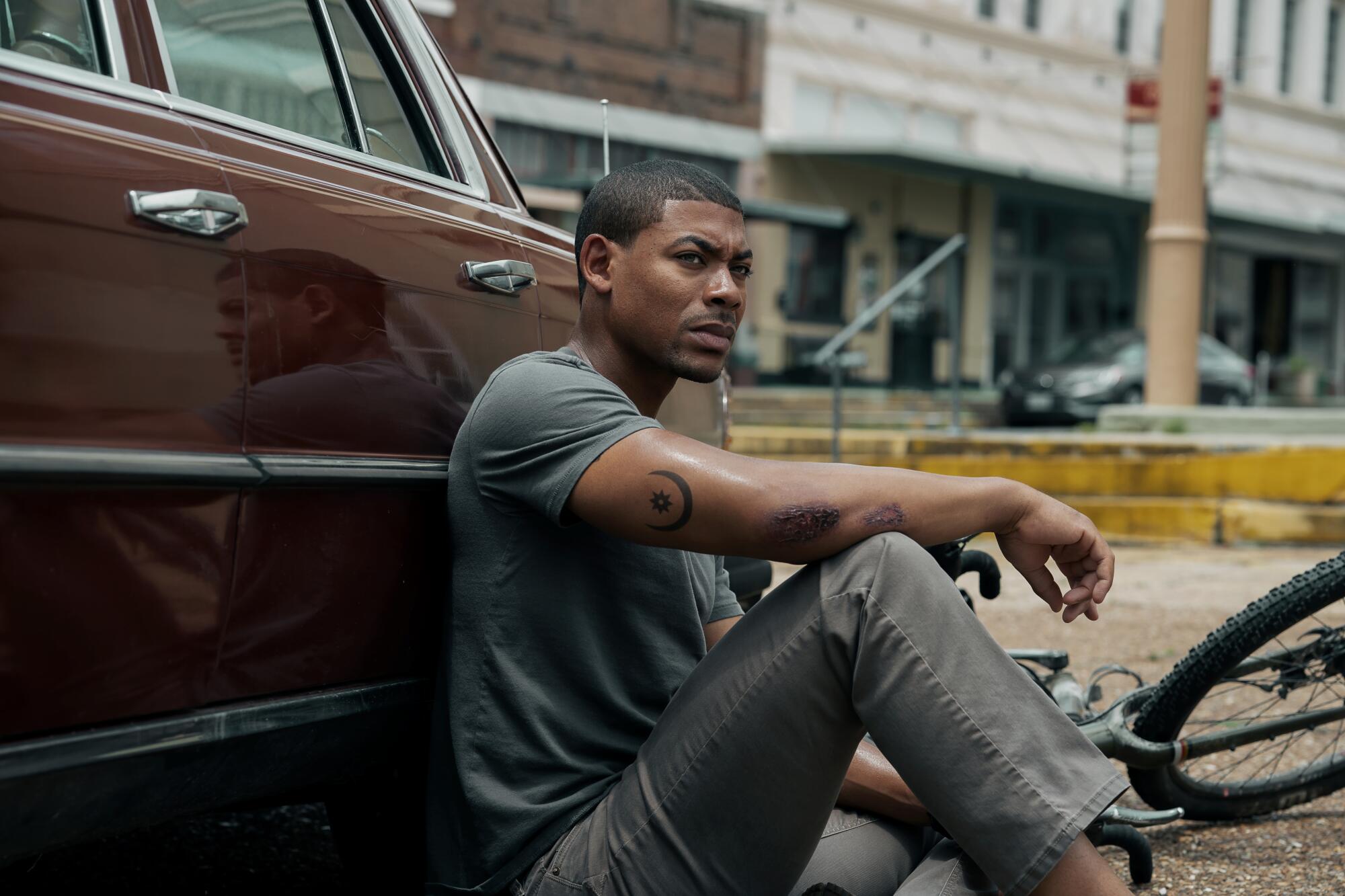With a bicycle lying on the ground behind him, a man in a T-shirt and pants sits against an old sedan.