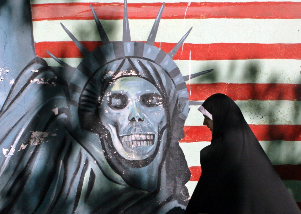 A veiled woman walks past graffiti of a skull-like Statue of Liberty on a wall painted with red and white stripes.