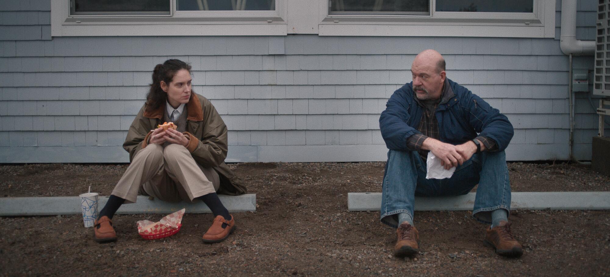 A woman eats a sandwich sitting next to a man in a parking lot.