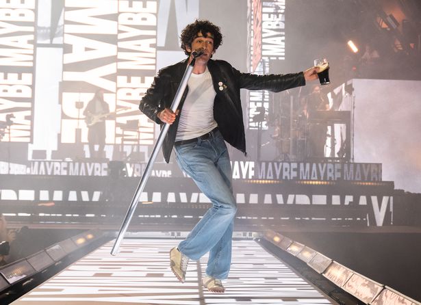 Matty Healy in a white top, black jacket and jeans on stage at Glastonbury.