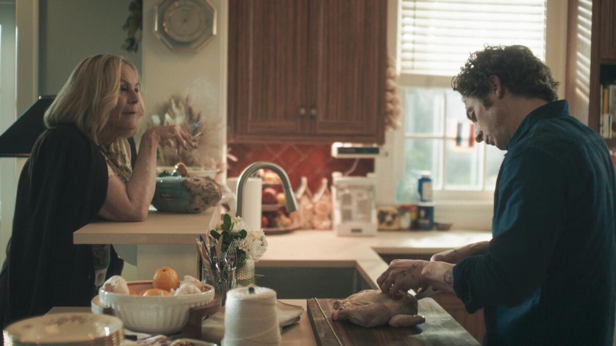 A woman stands at kitchen bar and looks at a man trussing a raw chicken.
