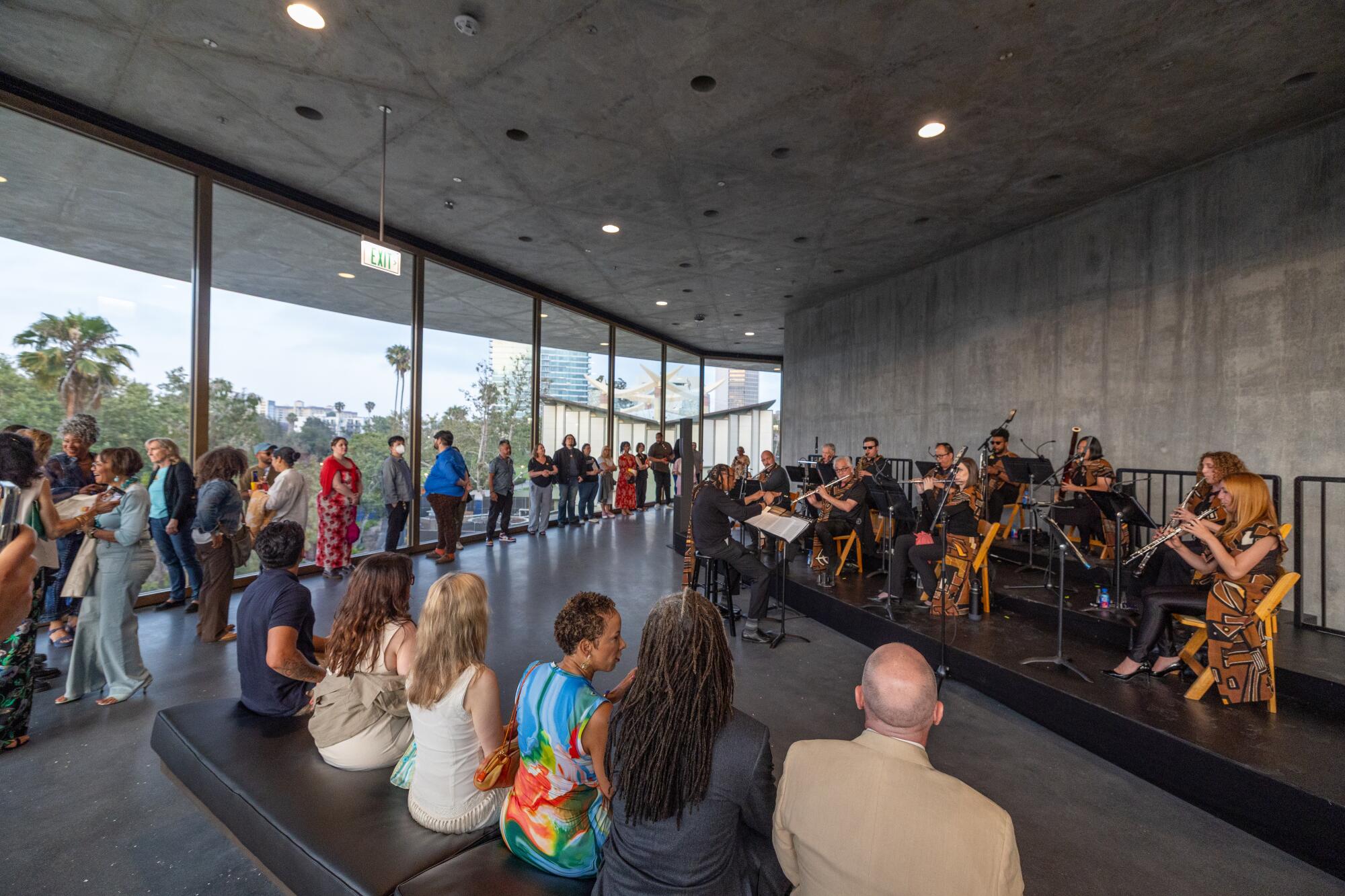 Musicians perform against the stark concrete walls of the David Geffen Galleries, as visitors stand along a wall of glass.