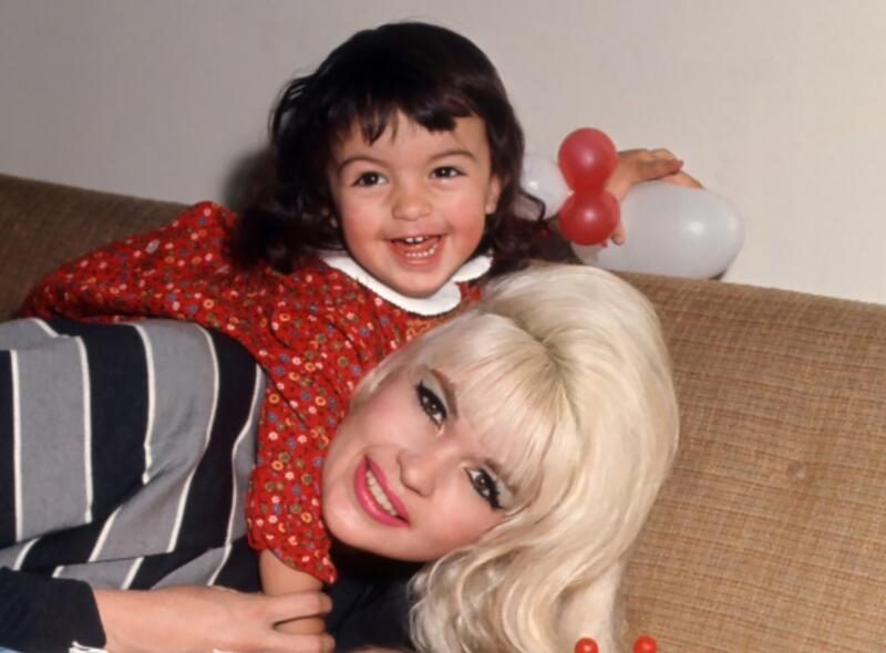 A young girl hangs on top of a woman who is lying down on a couch. 