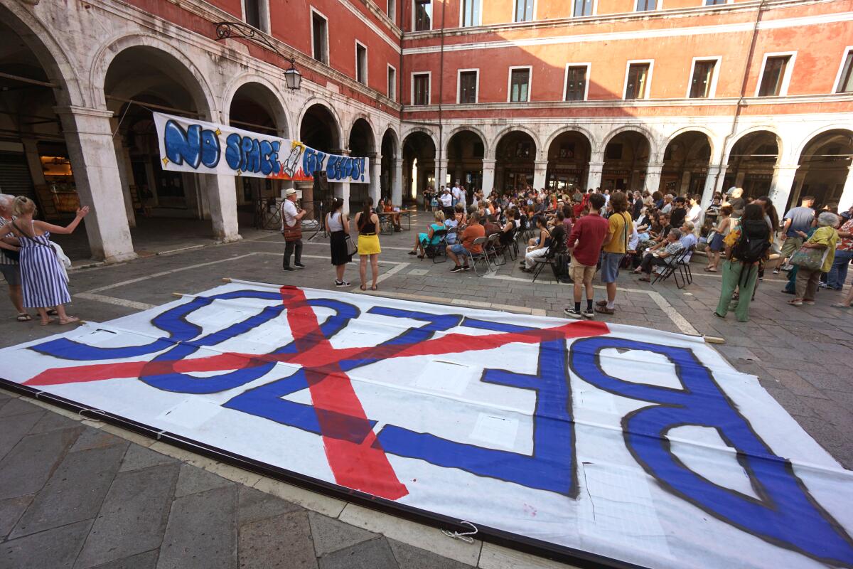 A massive banner reading "Bezos" with an X over it lies on the ground in front of a seated group of Venice residents