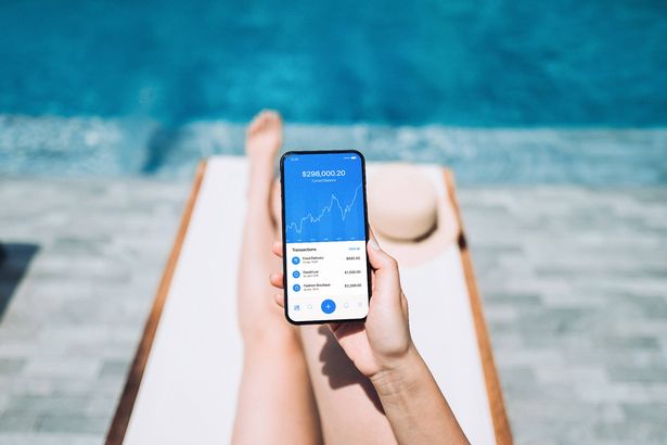 Low section of young woman managing online banking with mobile app on smartphone while enjoying the sun on lounge chair by pool side. Tracking and planning spending. Transferring money, paying bills, checking account balance. Smart banking with technology