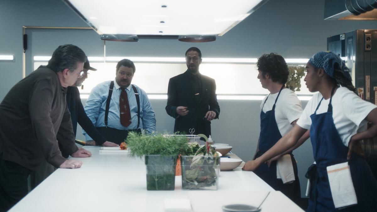 A group of men and a woman stand around a kitchen prep table with leafy greens and bowls on one side.