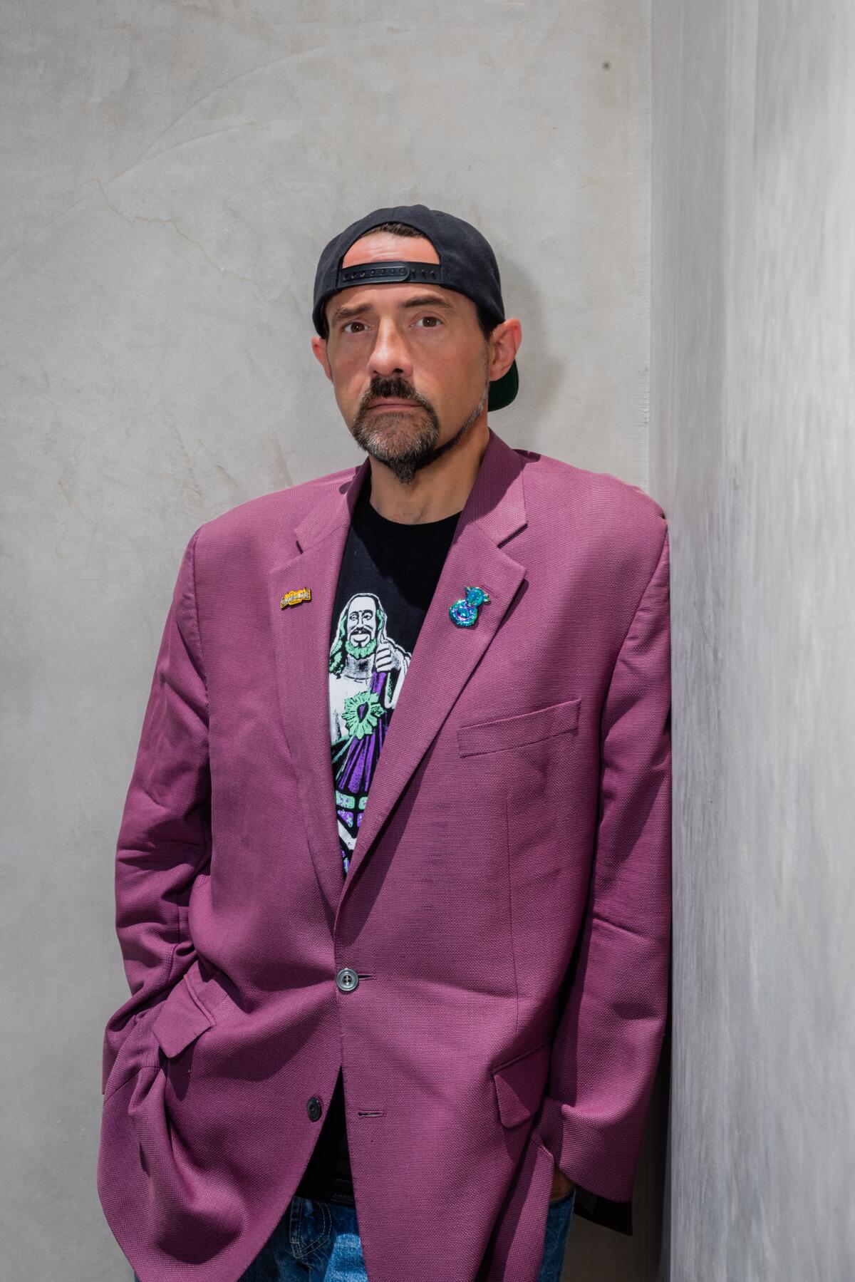 Kevin Smith, in a purple blazer and backwards cap, leans against a wall. 