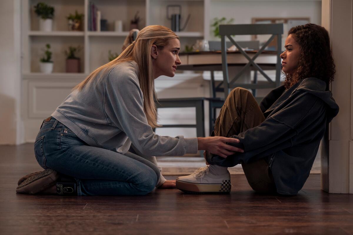 A woman in a blue top and jeans kneels in front of a teenage girl in a hoodie and brown pants.