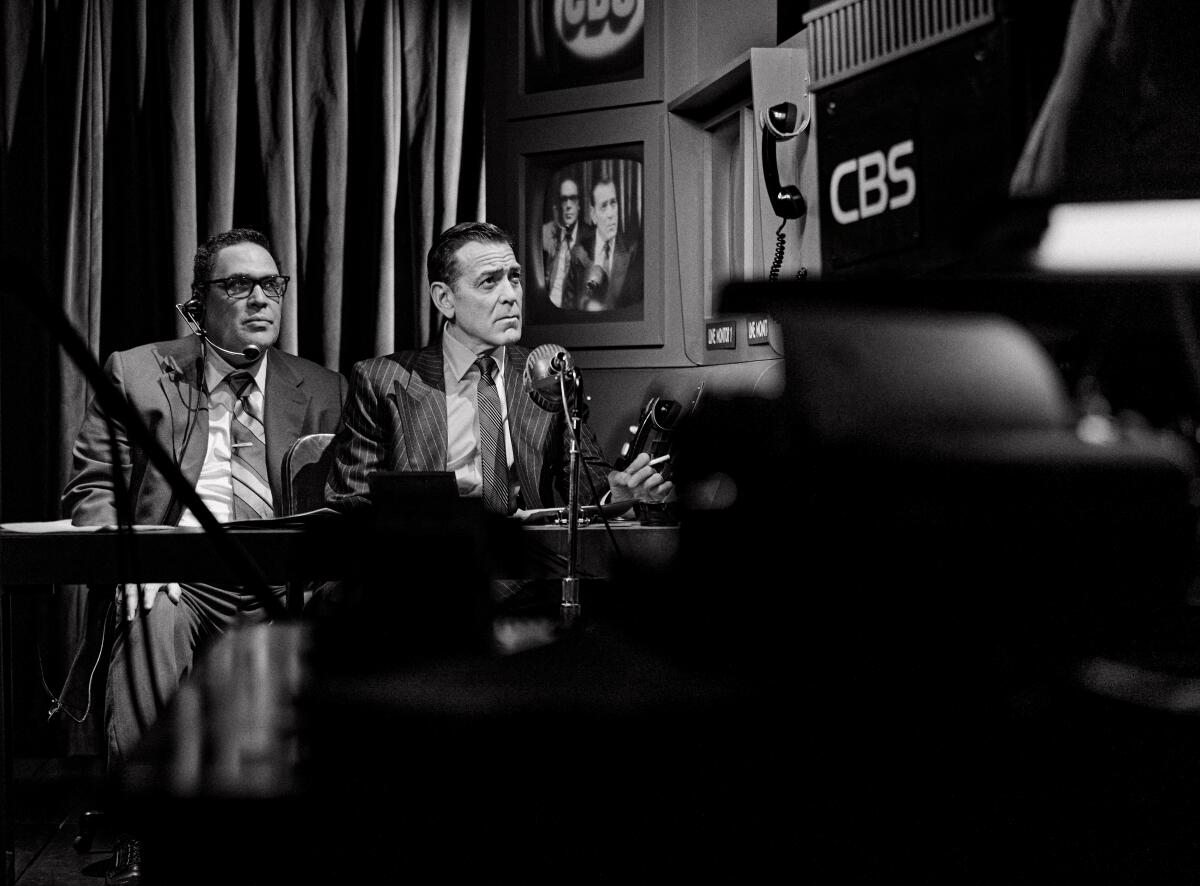 Two men in suits sit behind a desk with microphones. Screens are seen behind them.