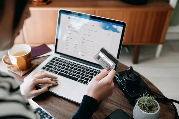 Over the shoulder view of young Asian woman shopping online for flight tickets on airline website with laptop, entering credit card details to make mobile payment at home. Camera and passport on the table. Travel planning. Booking a holiday online