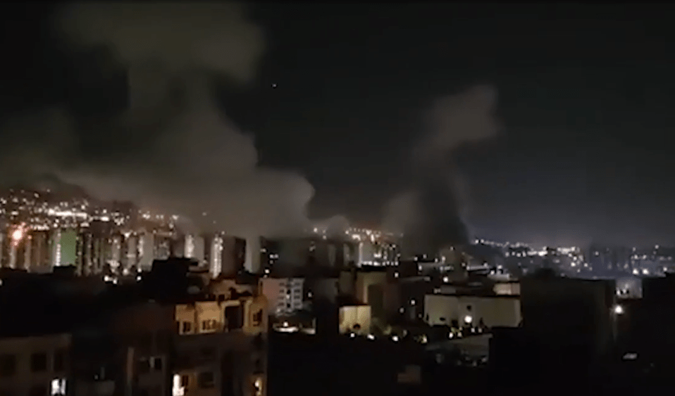 Nighttime view of a city with smoke and fire in the distance.