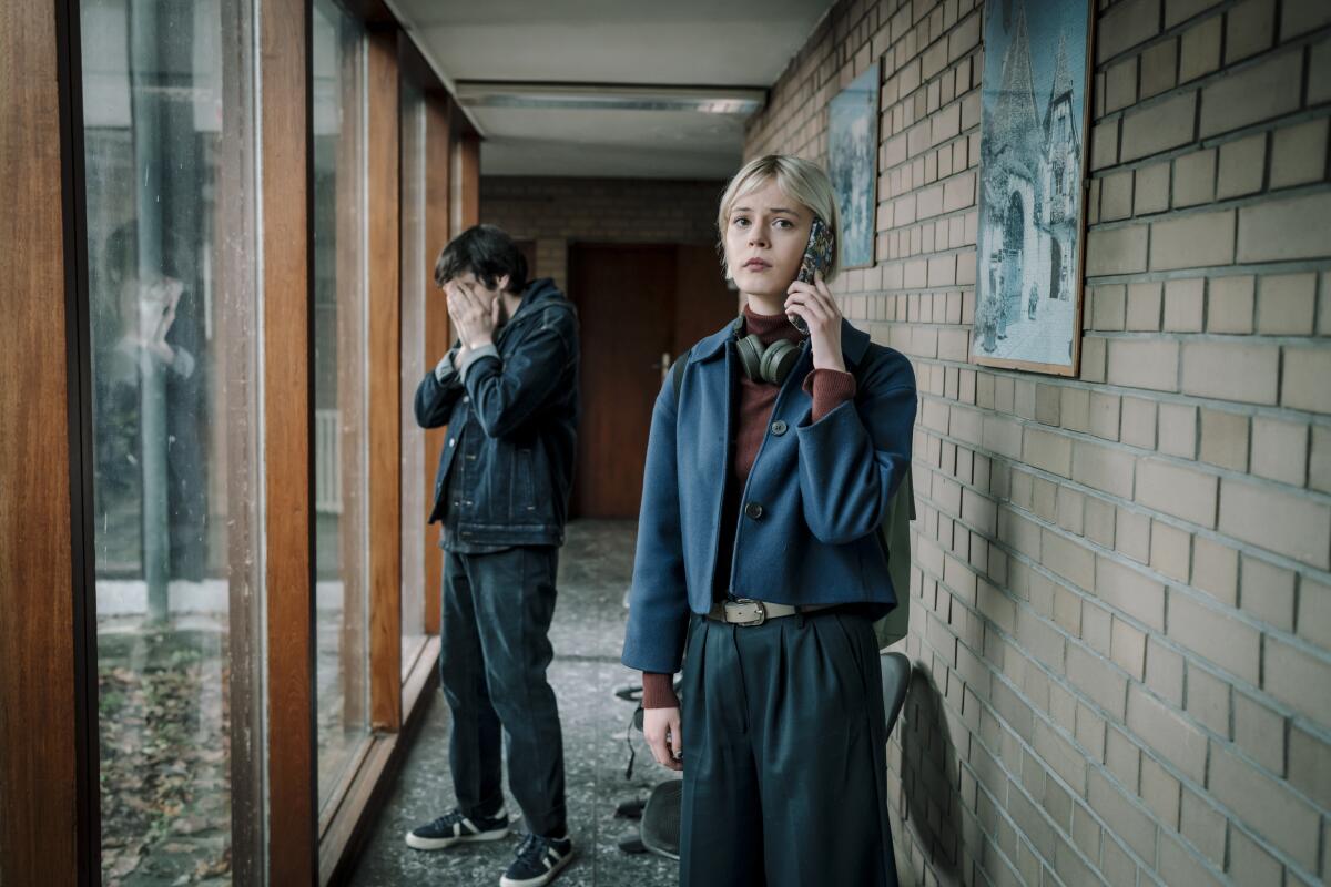 A man stands near a full-length glass window covering his face with his hands as a woman holds a cell phone up to her ear.