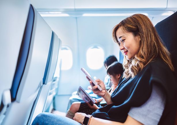 Young Asian woman consults her smartphone aboard an airplane, reviewing her travel plans. Her daughter is reading book next to her. Concept of family travel and vacation