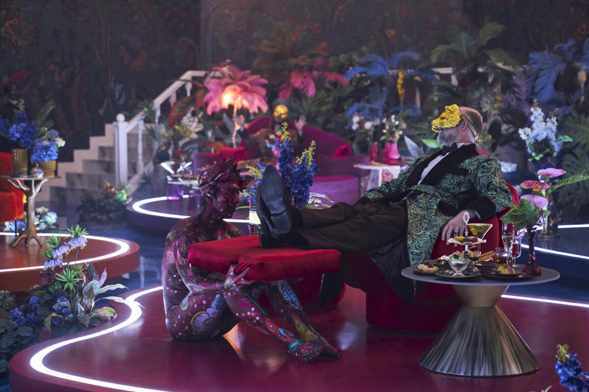 A man with a beard and golden mask lounging in a red chair with his feet propped up on a red cushion held by a person.