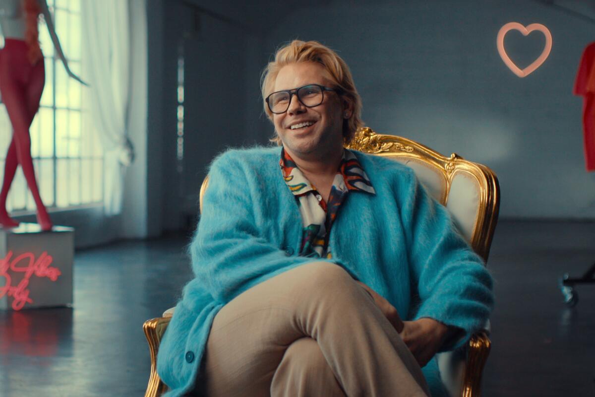 A smiling man in a fuzzy blue sweater and brown slacks sits a chair.