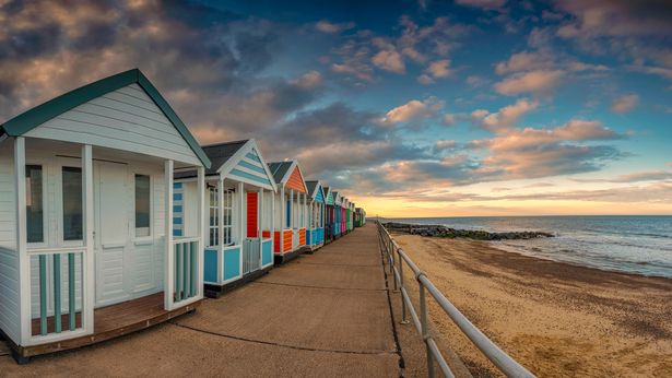 Image shows the scene along a beach path with some brightly coloured beach huts. The scene is calm, bright and warm but the light is fading as dusk is taking hold of the scene. There are no people in the scene.