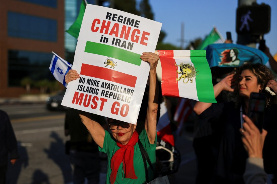 Protestor holding Israeli and pre-Islamic Republic Iranian flags and a sign that reads "Regime Change in Iran" and "No More Ayatollahs Islamic Republic Must Go".