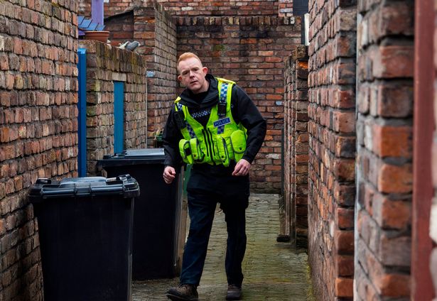 Colson Smith, in a police uniform, walking down an alley on the set of Corrie for a scene.
