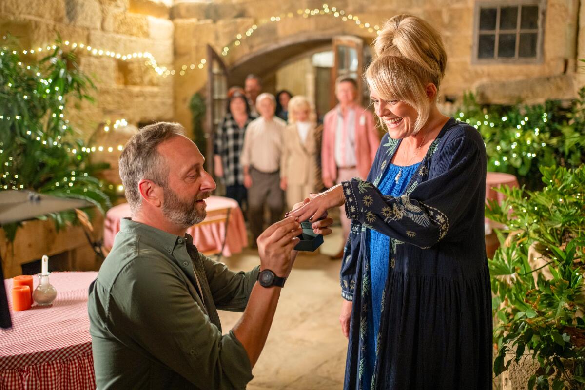 A kneeling man in a green shirt places a ring on the finger of a blond woman