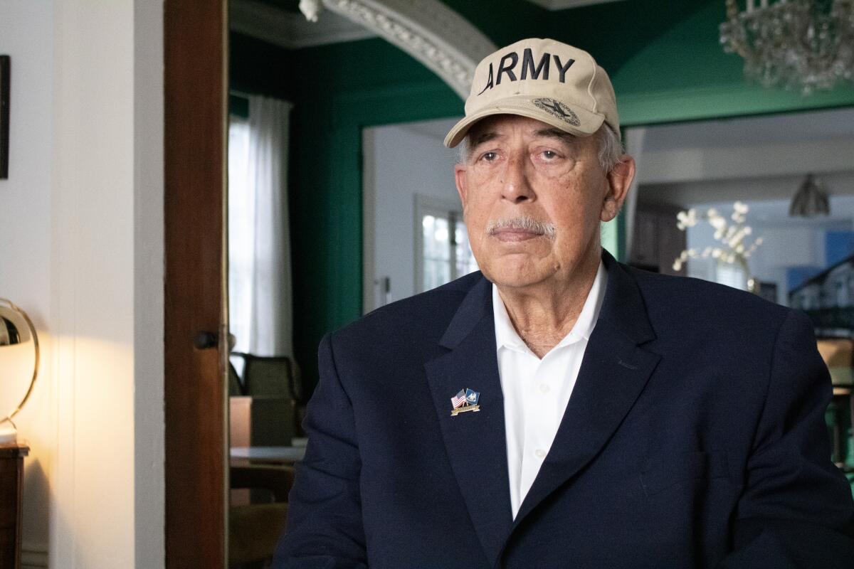 An older man in a white shirt and blue blazer wearing a ball cap that says Army.