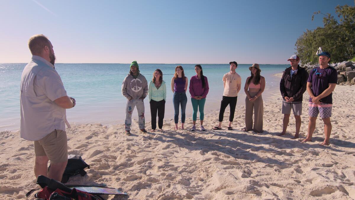 A man stands opposite a group of eight people standing on a beach near the water.