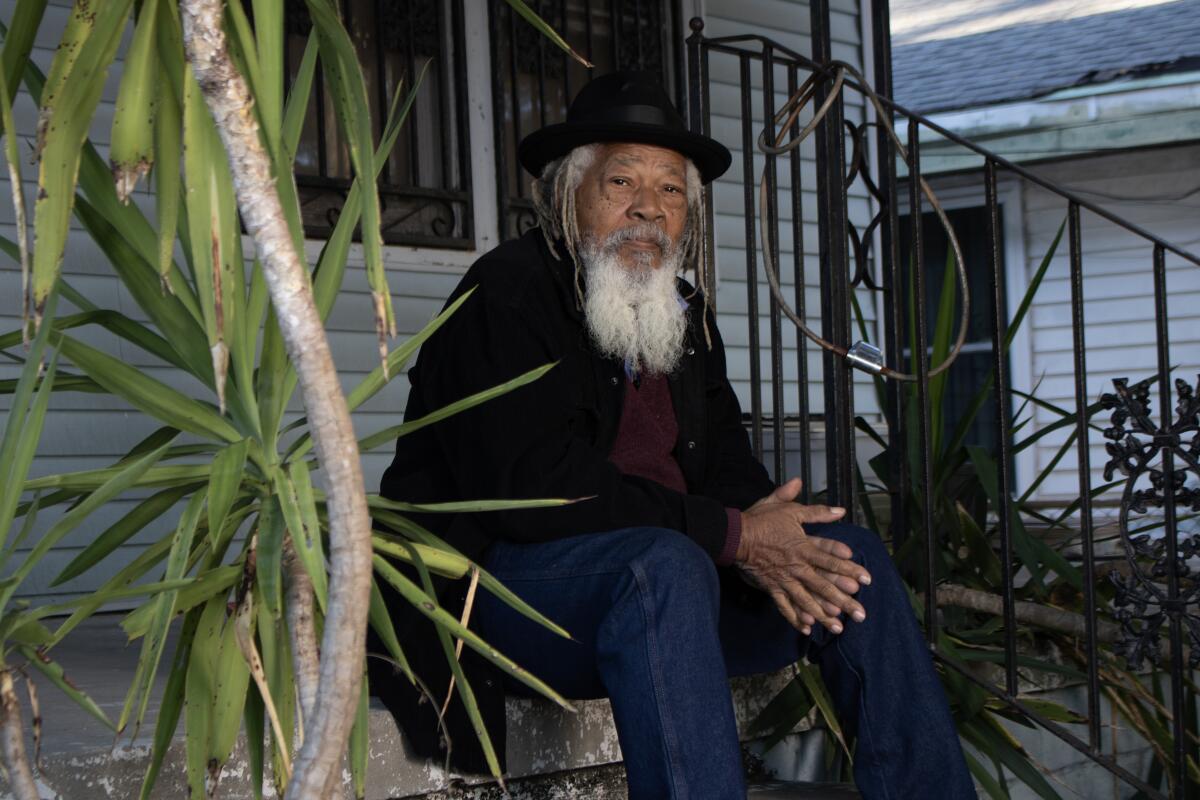 A man in black hat, dark jacket and jeans sits on the stoop of a house.