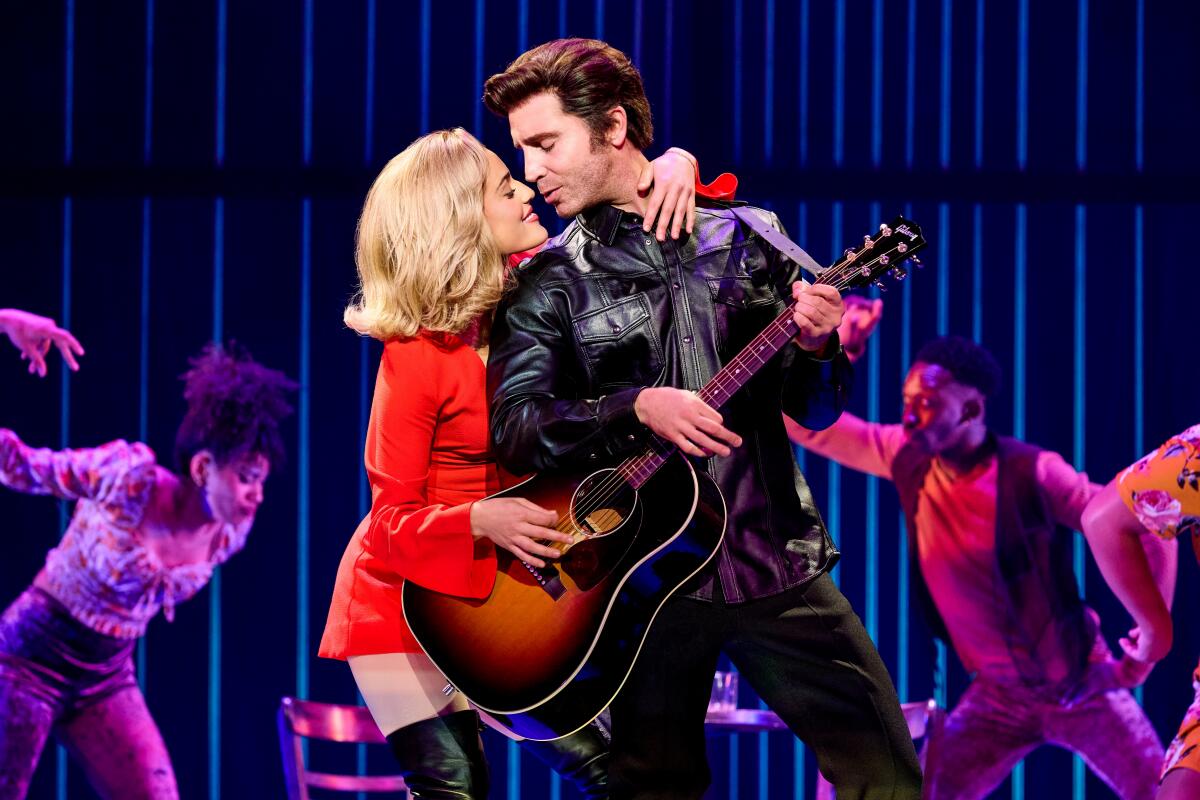 A woman in a red dress with her arm around a leather-jacked man with a guitar in a stage show.