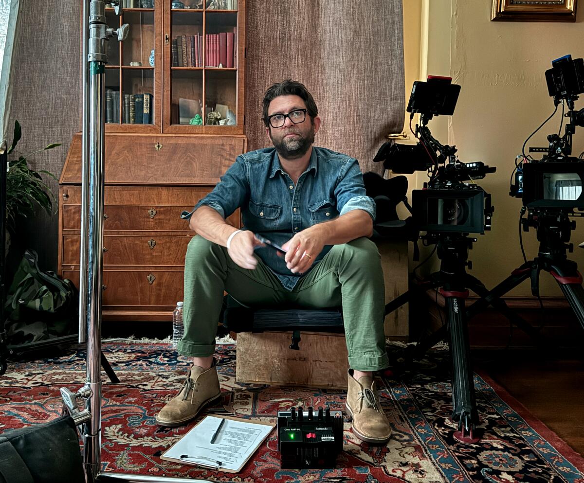 A man in a denim shirt and green pants sits on a chair with camera equipment around him.