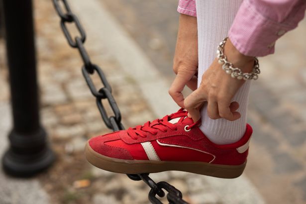 Moment of casual elegance captured as unseen adult woman, clad in chic outfit, wearing delicate pearl bracelet and metallic bangle on wrist, adding touch of sparkle to sporty aesthetic, tightening laces of vibrant red sneaker while resting foot on city street chain-link fence.