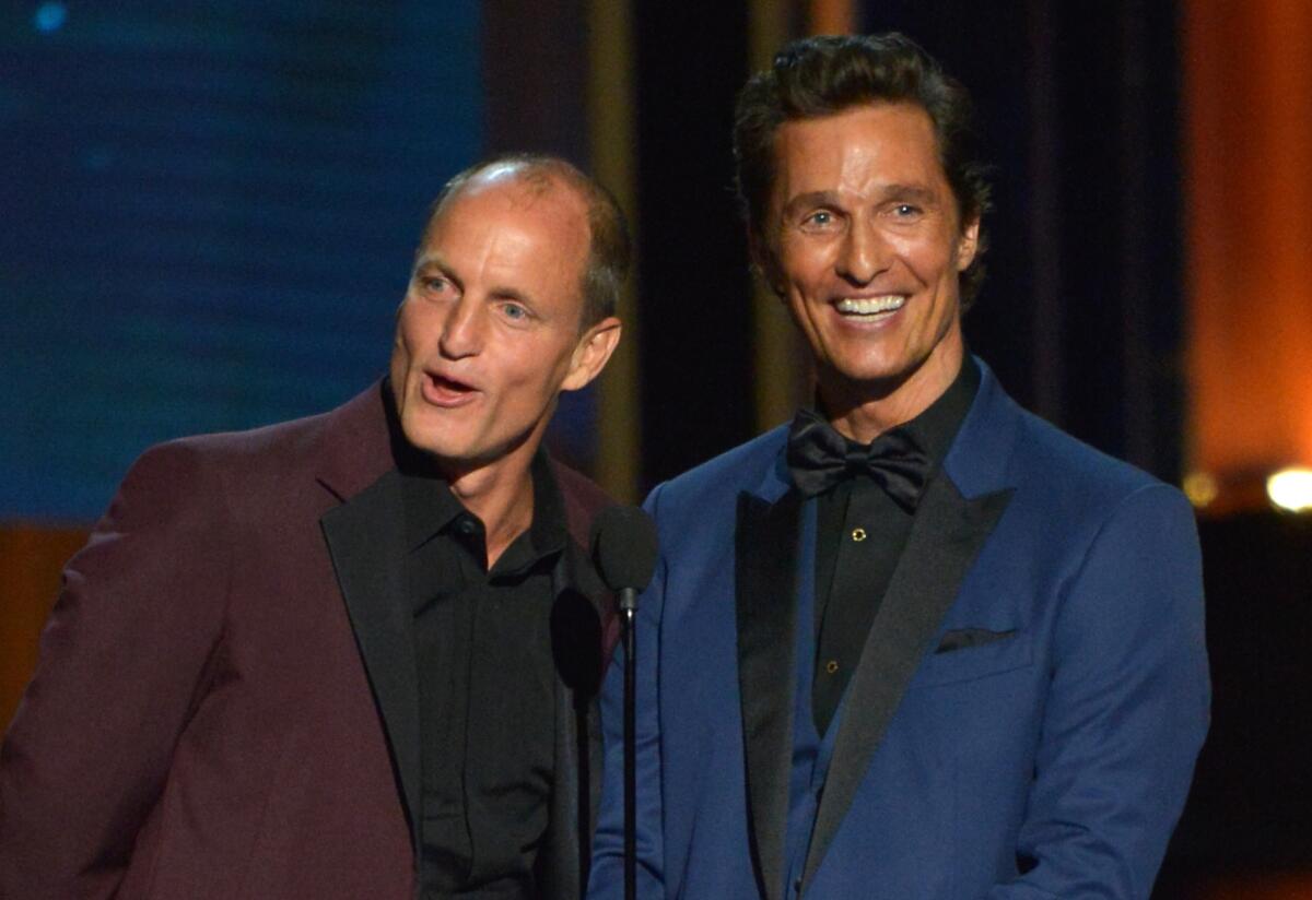 Woody Harrelson wearing a maroon suit and Matthew McConaughey wearing a blue suit smiling on stage