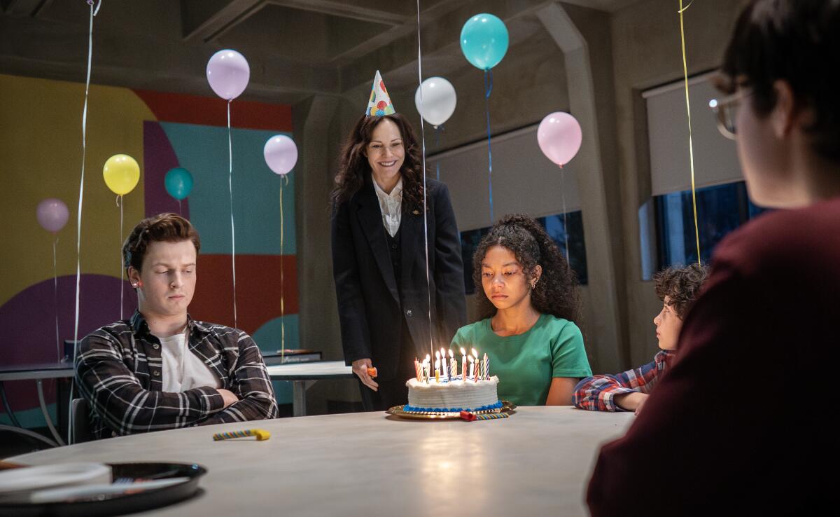 A group of children sit at a table around a birthday cake as a woman stands behind them.