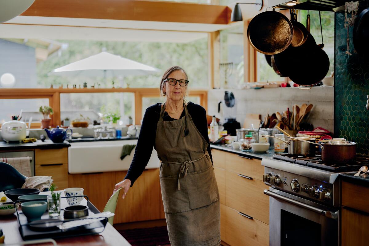 A woman stands in an apron in a kitchen with kitchenware and pots around her. 