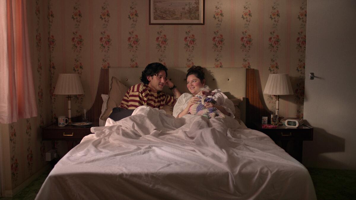 A man and a woman with a small dog on her lap lay in bed looking at one another.