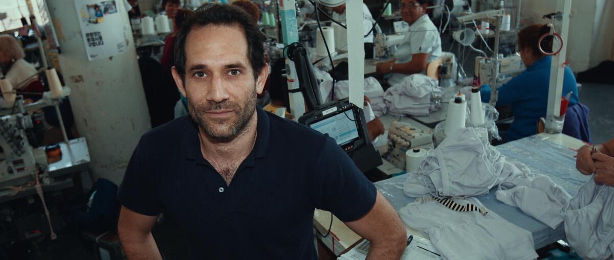 A man in blue polo shirt stands in a warehouse where women sewing clothing are seen in the background.