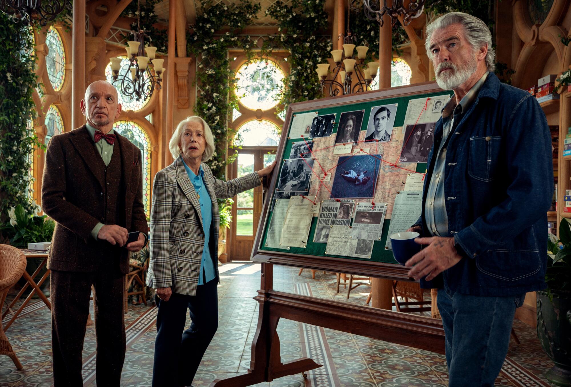 Three older people standing around a board covered in photos, newspaper clippings and a map.