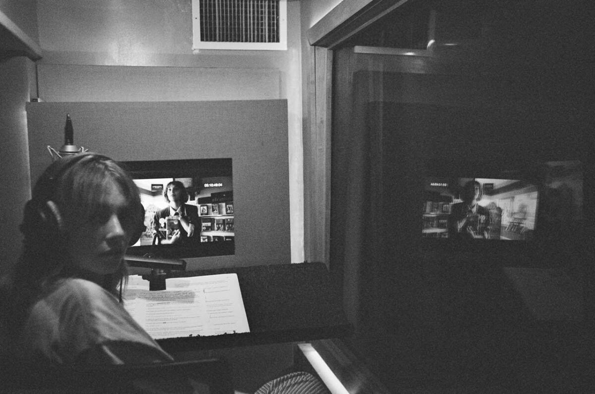 A woman in front of a microphone and a monitor records a narration.