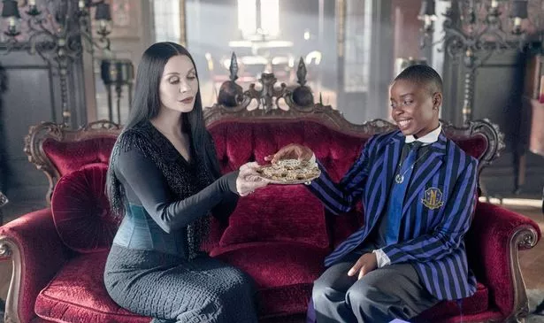 A woman offers a girl a plate of food as they sit on a couch