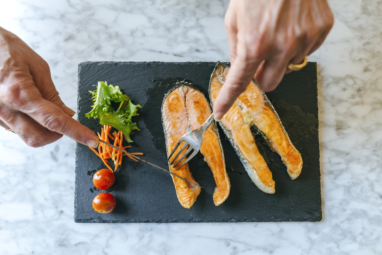 Close-up of woman's hands cutting grilled salmon fillets on plate with salad