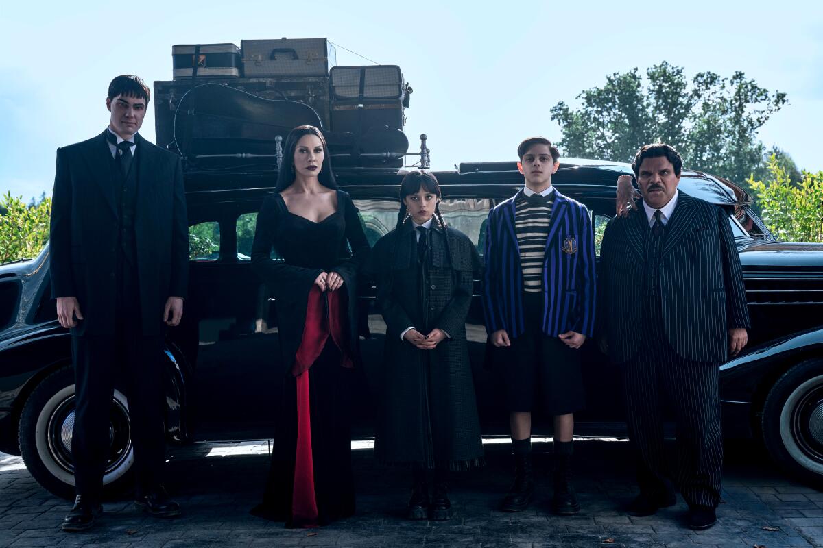 A group of non-smiling people stand in front of a vintage black car.