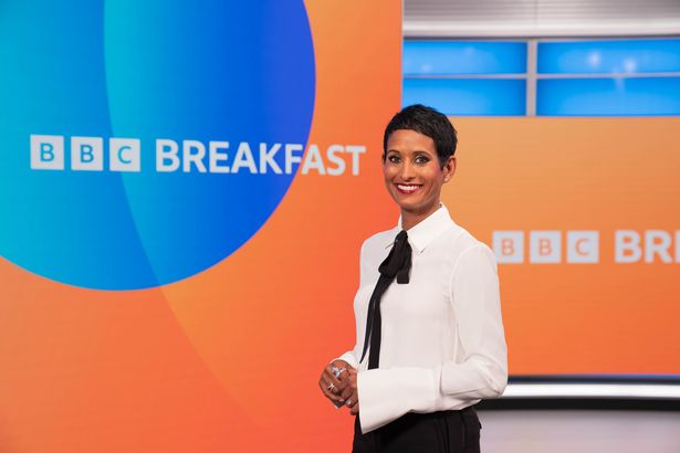 Photo shows a smiling Naga Munchetty dressed in a white blouse and black trousers stood in front of a screen with the BBC Breakfast logo on